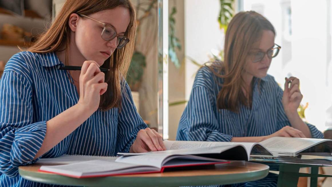 A young female writer is working on an assignment. Her reflection shows in a mirror next to her.