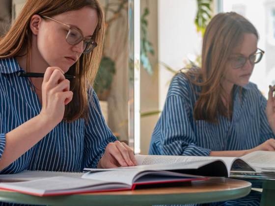 A young female writer is working on an assignment. Her reflection shows in a mirror next to her.