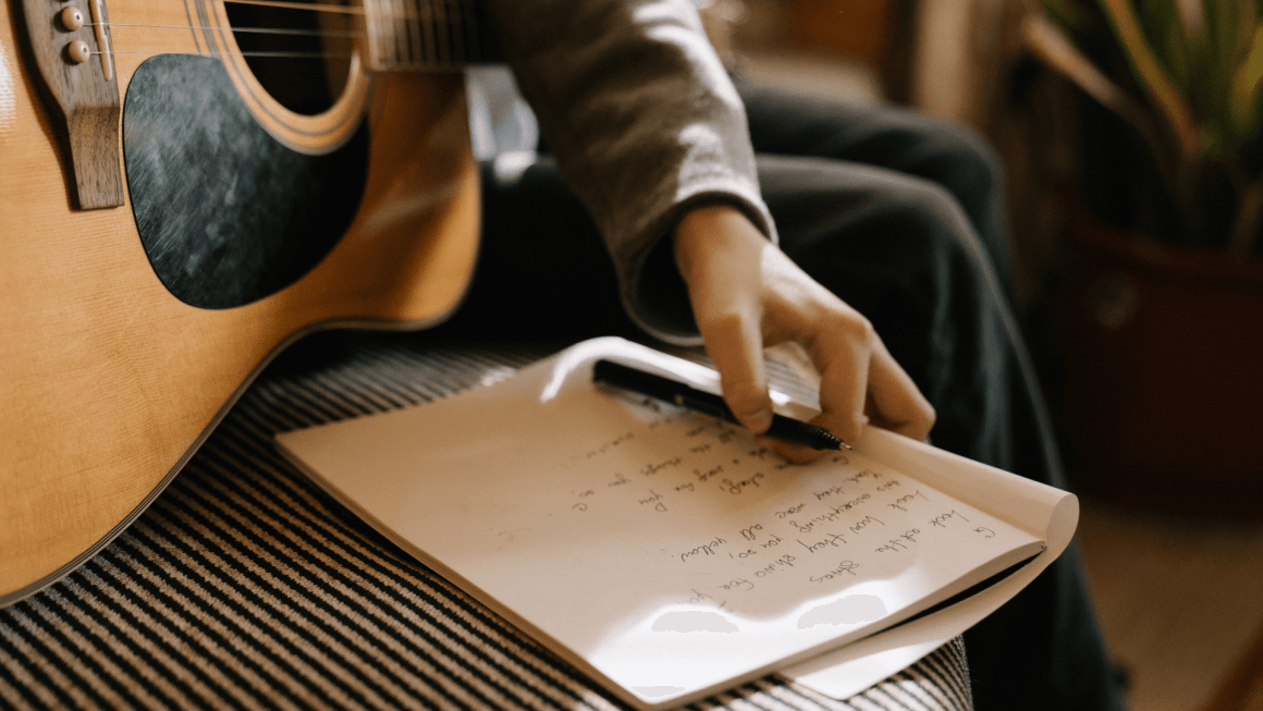 A hand is seen holding a notebook with song lyrics, next to a guitar.