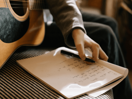 A hand is seen holding a notebook with song lyrics, next to a guitar.