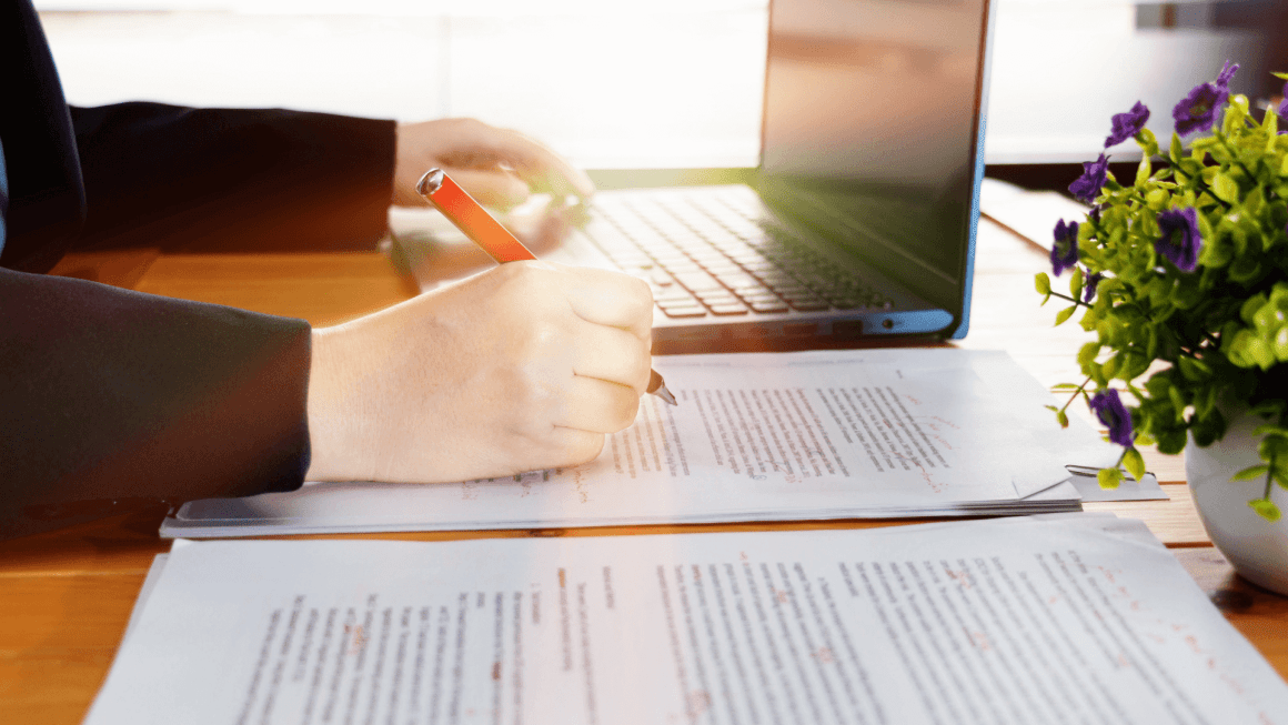 Woman with red pen marking up a research paper, next to a laptop