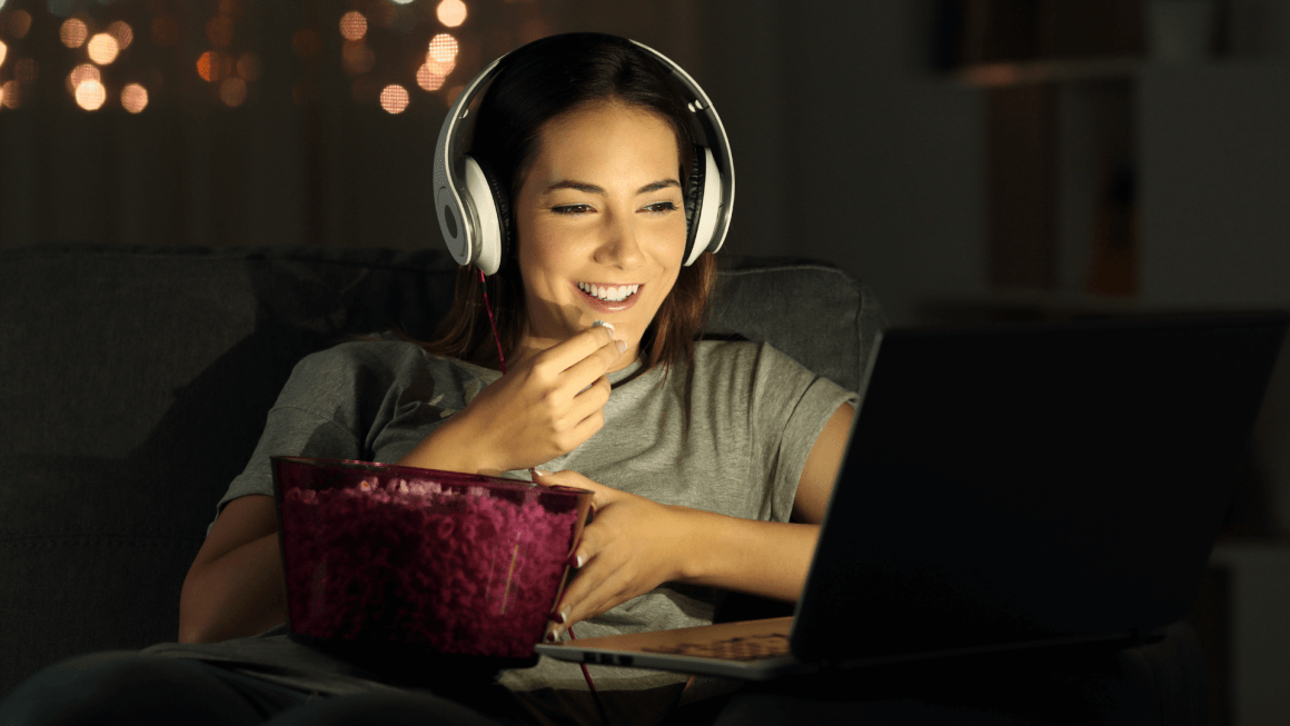 Woman wearing headphones and eating popcorn while watching a movie on her laptop.