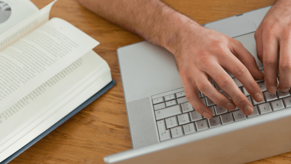 hands typing on a keyboard with an open book nearby.