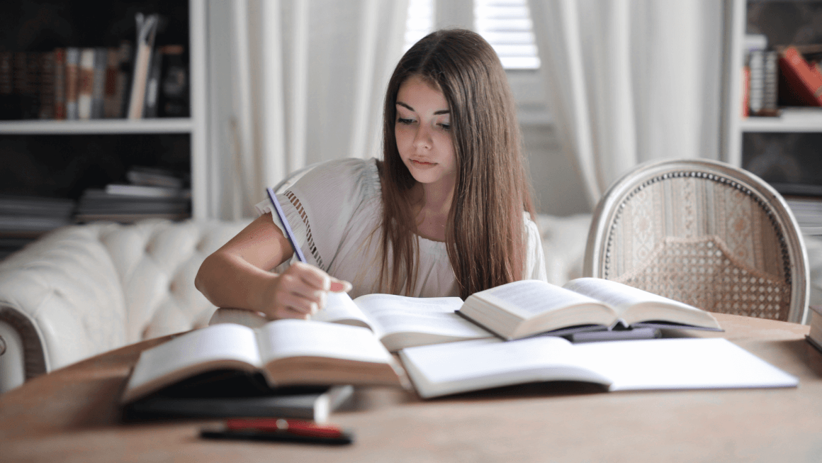 Young woman sitting at a table writing with many resource books open nearby