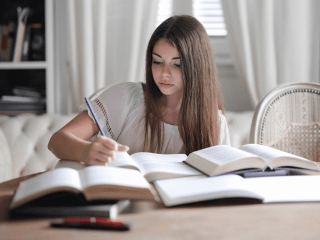 Young woman sitting at a table writing with many resource books open nearby