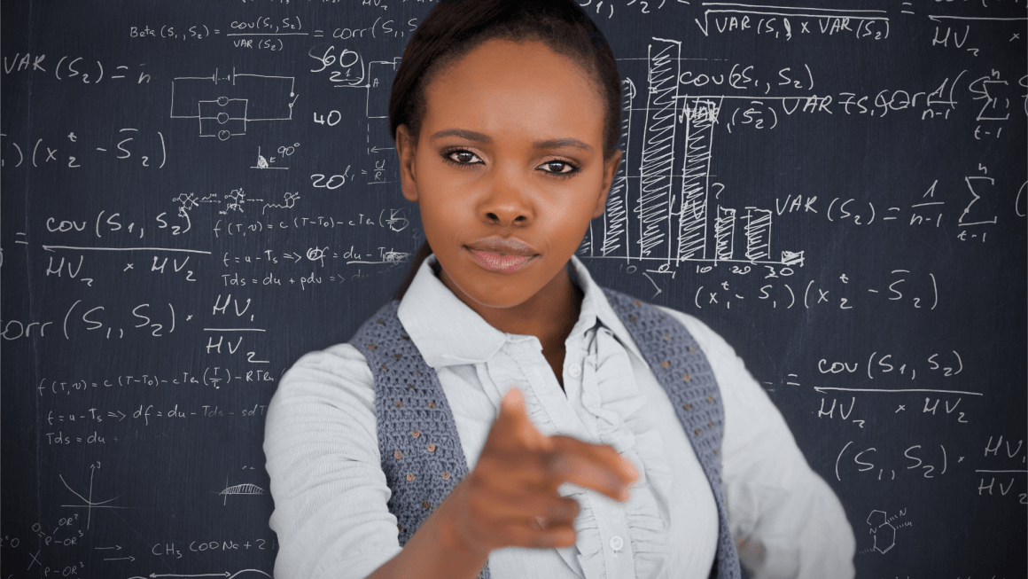 Teacher standing in front of a blackboard, pointing at the camera